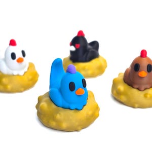 Könnte beinhalten: Vier farbenfrohe Hühnerfiguren, jede auf einem gelben, strukturierten Sockel. Die Hühner sind weiß, schwarz, braun und blau, mit roten Kämmen und orangen Schnäbeln. Die Figuren sind vor weißem Hintergrund.