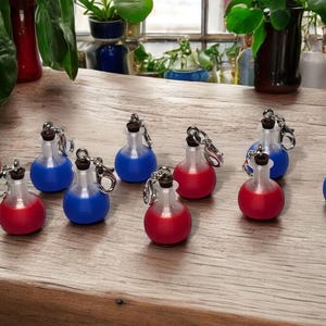 May include: Ten small glass bottles with silver caps and keychains. The bottles are filled with red and blue liquid. The bottles are arranged on a wooden surface.