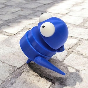 May include: A blue 3D printed toy with large white eyes and a segmented body. The toy is standing on a gray stone surface.