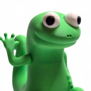 May include: A bright green, 3D-printed gecko figurine with large, expressive eyes and a friendly wave. The gecko has a smooth texture and is set against a plain white background. The figurine is a whimsical decorative item.
