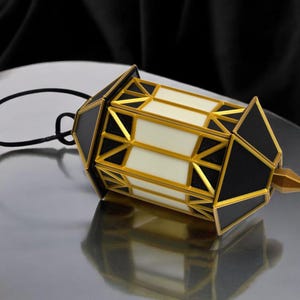 Puede incluir: Una linterna decorativa con diseño geométrico en negro, dorado y blanco. La linterna tiene un lazo negro para colgar y un remate dorado. Descansa sobre una superficie reflectante. El diseño incorpora triángulos y rectángulos.