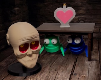 May include: A white ceramic head with red eyes and a light brown face, a green and blue creature with large eyes, and a heart-shaped bottle with pink liquid on a wooden table.