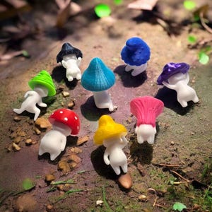 Puede incluir: Un conjunto de figuras de hongos con sombreros coloridos en rojo, amarillo, verde, azul y morado. Las figuras, con cuerpos blancos, están dispuestas sobre una superficie natural con piedras y follaje, creando una escena boscosa.