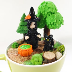May include: A miniature witch figurine in a black dress and pointed hat stands next to a cauldron filled with green liquid. A black cat figurine sits beside a small tree. The scene is set in a green teacup with brown sand and green moss.