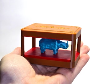 May include: A small, brown and red rectangular display case with a blue rhinoceros figurine inside. The case is held in a person's hand against a white background. The figurine is a solid color and has a textured surface.