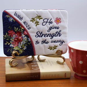 May include: A quilted square with floral embroidery and the text "He gives Strength to the weary" is displayed on a gold stand. A red and white polka-dot teacup with the word "Love" is next to it.