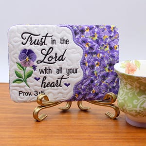 Può includere: Targa ricamata decorativa con la scritta "Trust in the Lord with all your heart" e un motivo floreale, esposta su un supporto dorato. Accanto, una tazza da tè a fantasia verde e bianca con un motivo floreale.