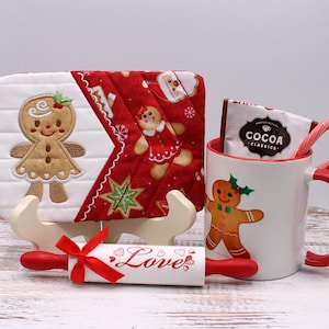 May include: A festive holiday gift set featuring a gingerbread-themed mug, a quilted pot holder, a rolling pin with "Love" text, and cocoa mix. The mug and pot holder have gingerbread man designs.