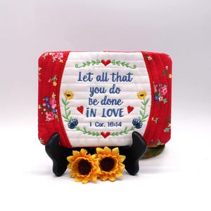 Può includere: Un oggetto in tessuto trapuntato rosso e bianco con il testo ricamato "Let all that you do be done IN LOVE" e dettagli floreali. L'oggetto è esposto su un supporto nero, con due girasoli gialli alla base.