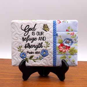 Può includere: Un quadrato decorativo con il ricamo "Dio è il nostro rifugio e la nostra forza" e Salmo 46:1. Il quadrato presenta uno sfondo bianco trapuntato, ricami floreali e una sezione a motivi floreali blu. È ricamata anche una farfalla gialla.