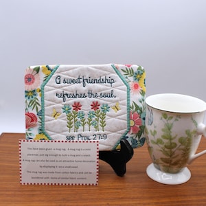 Pode incluir: Um tapete floral para caneca com o texto "A sweet friendship refreshes the soul" e "see Prov. 27:9". Uma caneca de cerâmica branca com um design floral está ao lado do tapete. O tapete para caneca está em um pequeno cavalete preto.
