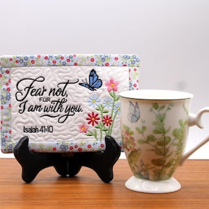 Pode incluir: Uma caneca de cerâmica branca com um design floral e borboletas, ao lado de um quadrado acolchoado com o texto "Fear not, for I am with you." O quadrado é suportado por um suporte preto. A caneca tem uma alça curva e uma base branca.