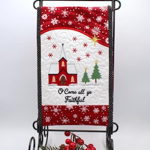 May include: A Christmas-themed quilted wall hanging featuring a red church, snowflakes, and the text "O Come all ye Faithful." The design is set against a red and white checkered background, displayed on a black metal stand.