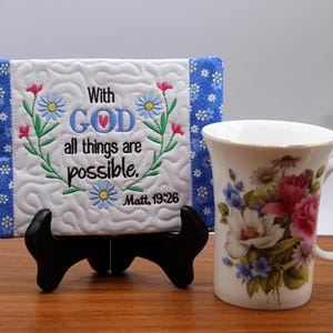 Può includere: Targa trapuntata bianca con il testo ricamato "With GOD all things are possible" e un motivo floreale, esposta su un supporto nero. Una tazza da tè decorata con fiori con bordo dorato è accanto alla targa.