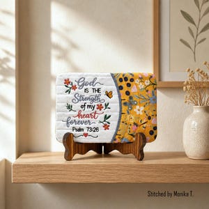 Può includere: Un cartello rettangolare in tessuto trapuntato su un supporto in legno scuro. Il lato sinistro è bianco con il testo ricamato God IS THE Strength of my heart forever Psalm 73:26, decorato con piccoli fiori e una farfalla. Il lato destro è giallo.