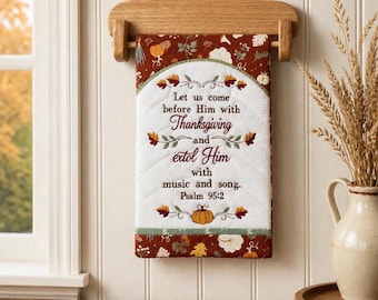May include: A rectangular quilted wall hanging featuring a white centre panel with embroidered text reading Let us come before Him with Thanksgiving and extol Him with music and song. Psalm 95:2, surrounded by a rust-coloured border with autumn leaves.