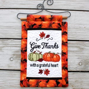 Op de afbeelding: Een decoratieve wandhanger met herfstthema met een metalen hanger. De stof heeft een pompoenpatroon en een centraal paneel met de woorden "Give Thanks with a grateful heart" en geborduurde pompoenen en bladeren.