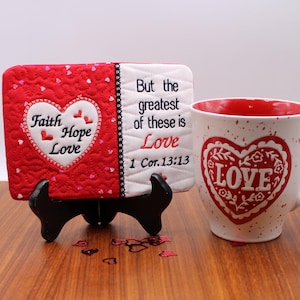 Può includere: Una tazza e una targa decorativa a tema cuore, rosse e bianche. La tazza presenta un cuore rosso con la scritta "LOVE". La targa mostra "Faith, Hope, Love" e "But the greatest of these is Love 1 Cor. 13:13".
