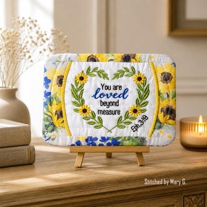 Può includere: Un cartello rettangolare in tessuto trapuntato su un piccolo cavalletto di legno con stampa di girasoli e fiori blu. Il centro presenta una ghirlanda ricamata a forma di cuore con il testo You are loved beyond measure Eph.3:19.