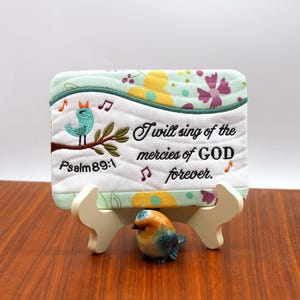 Könnte beinhalten: Dekorative, gesteppte Plakette mit dem Text "I will sing of the mercies of GOD forever." und "Psalm 89:1." Die Plakette zeigt einen türkisfarbenen Vogel auf einem Ast und Noten. Vor der Plakette sitzt eine kleine Keramik-Vogelfigur.