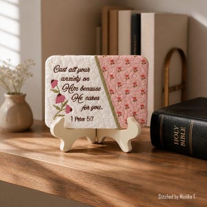 May include: A decorative, rectangular piece with embroidered text and floral accents, displayed on a white wooden stand. The text reads "Cast all your anxiety on Him because He cares for you. 1 Peter 5:7." The background is a mix of white and pink floral fabric.