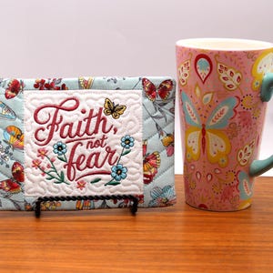 Può includere: Una placca decorativa trapuntata con la scritta ricamata "Faith, not fear", con motivi floreali e farfalle. Accanto, una tazza in ceramica rosa con manico blu e motivo a farfalle. L'interno della tazza è bianco.
