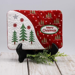 May include: A quilted Christmas coaster with a white and red design. The white section features embroidered green trees, snowflakes, and a star. The red section has a repeating tree pattern and the text "Merry Christmas".