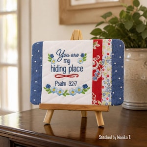 Può includere: Un cartello rettangolare in tessuto trapuntato esposto su un piccolo cavalletto di legno. Il pannello centrale bianco presenta un testo ricamato in blu che recita You are my hiding place Psalm 32:7, incorniciato da motivi floreali blu e verdi. I lati sono in tessuto blu a pois.