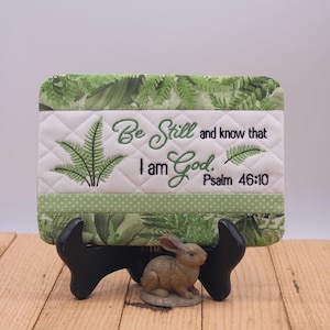 Può includere: Un pezzo decorativo trapuntato con il testo ricamato "Fermatevi e sappiate che io sono Dio. Salmo 46:10" e disegni di foglie di felce. Il pezzo è sostenuto da un supporto nero, con una piccola statuina di coniglio davanti.