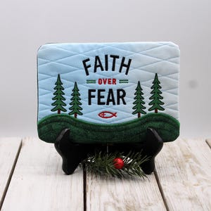 Può includere: Targa decorativa trapuntata azzurra con le parole "FAITH OVER FEAR" ricamate in nero. Il design include alberi verdi, una collina verde e un simbolo di pesce rosso. La targa è esposta su un supporto nero.