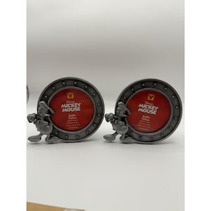 May include: Two round, silver-colored photo frames with a Mickey Mouse design. Each frame has a red interior and a decorative border. The frames are designed to hold 3x3 inch photos, or 7x7 cm.