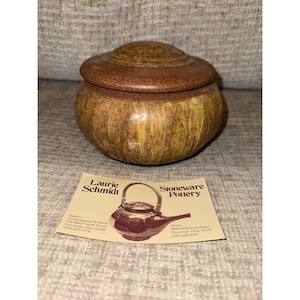 Peut inclure: Un pot en grès fait à la main avec un couvercle. Le pot présente des tons bruns et jaunes, avec une surface texturée. Le couvercle est brun foncé. Une carte de visite avec "Laurie Schmidt" et "Stoneware Pottery" est visible.