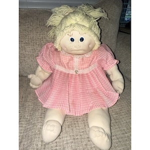 Könnte beinhalten: Eine weiche Stoffpuppe mit blasser Haut, blauen Augen und Sommersprossen, die hellgelbes Wollhaar in einem unordentlichen Dutt trägt. Die Puppe trägt ein rosa-weiß kariertes Kleid mit kurzen Puffärmeln, einem Spitzenkragen und einer kleinen Schleife.