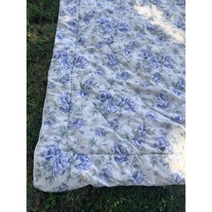 May include: A quilted blanket featuring a repeating pattern of blue roses and green leaves on a light cream background. The fabric has a stitched border and a soft, textured surface with visible quilting lines throughout the entire piece.