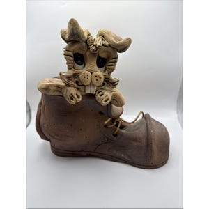 May include: A whimsical ceramic sculpture of a rabbit peeking out of a brown boot. The rabbit has large eyes and prominent teeth. The boot has laces and a worn appearance, suggesting a rustic or vintage aesthetic.