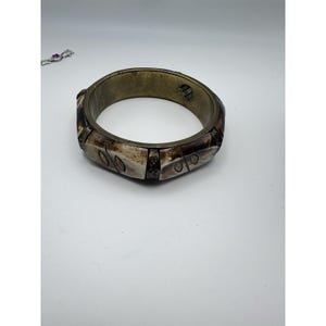 May include: A wide, antique-style bracelet with a brass-colored band and carved bone-like segments. The bracelet features geometric designs and a weathered appearance, suggesting age and craftsmanship. The bracelet is a statement piece of jewelry.