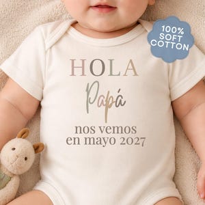 May include: A cream-colored short-sleeve baby bodysuit featuring the text "HOLA Papá nos vemos en mayo 2027" in a gradient brown font. A blue badge in the upper right corner displays the text "100% SOFT COTTON".