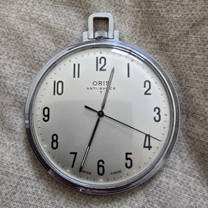 Op de afbeelding: Een zilverkleurig zakhorloge met een witte wijzerplaat en zwarte cijfers. De wijzerplaat toont de merknaam "ORIS" en de tekst "ANTI-SHOCK T". Het horloge heeft een lus aan de bovenkant om een ketting aan te bevestigen.