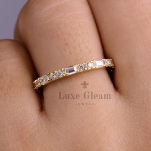 May include: A gold band ring with a series of small, clear gemstones. The ring features a mix of round and baguette-cut stones. The ring is on a hand. The Luxe Gleam Jewels logo is visible.