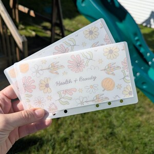 May include: Two translucent plastic cash envelopes for a binder, featuring a repeating pattern of pale yellow, pink, and orange flowers and leaves. The front envelope is clearly printed with the text Health + Beauty in a dark cursive font.