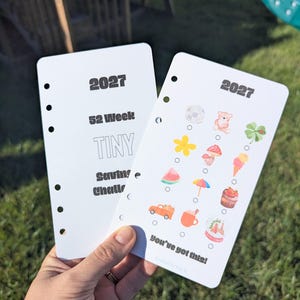 Könnte beinhalten: Zwei weiße Planereinlagen für eine 52-wöchige Spar-Challenge für 2027. Ein Blatt zeigt schwarzen Text, während das andere bunte Symbole wie einen Bären, einen Pilz, ein Eis und eine Wassermelone sowie den Text You've got this! am unteren Rand enthält.