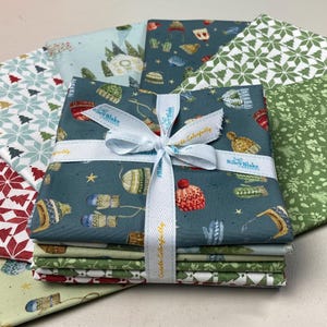 Może przedstawiać: Stos kwadratowych kawałków materiału o tematyce bożonarodzeniowej, związanych białą wstążką. Tkanina przedstawia wzory rękawic, czapek, płatków śniegu i drzew w odcieniach zieleni, czerwieni i niebieskiego. Na wstążce widnieją słowa "Riley Blake" i "Create Colorfully".