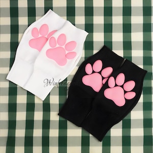 May include: Two pairs of socks, one white and one black, each featuring a pink paw print design. The socks are made of a soft material and are likely for pets or pet lovers. The background is a green and white checkered pattern.