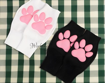 May include: Two pairs of socks, one white and one black, each featuring a pink paw print design. The socks are made of a soft material and are likely for pets or pet lovers. The background is a green and white checkered pattern.