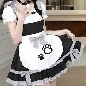 May include: Black and white maid dress with a ruffled skirt and a white apron featuring a paw print design. The dress has puff sleeves and is accented with bows. A black choker necklace completes the outfit.