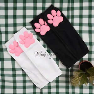 May include: Two pairs of long socks, one black and one white, each featuring a large pink paw print design near the top. The socks are displayed on a green and white checkered surface. The brand name "Winfredgoods" is printed on the white sock.