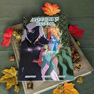 May include: A book titled "Anthia's Bloom" with a fantasy illustration on the cover. The cover art features several characters in shades of green, blue, and gold. The book is surrounded by autumn leaves and dice.