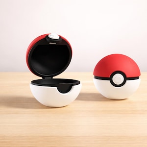 Pode incluir: Dois recipientes Poké Ball vermelhos, brancos e pretos, um aberto e outro fechado, estão sobre uma superfície de madeira clara. O recipiente aberto revela um interior preto. O recipiente fechado tem um botão branco.