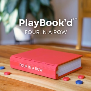 Könnte beinhalten: Ein rotes PlayBook'd-Spielcase mit dem Aufdruck "FOUR IN A ROW" auf der Seite. Das Spielcase ist geschlossen und liegt auf einer Holzoberfläche, mit roten und blauen Spielsteinen verstreut. Das Spiel ist für zwei Spieler konzipiert.
