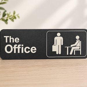Może przedstawiać: Czarny prostokątny znak z białymi słowami "The Office" i białą grafiką dwóch ludzików. Jedna postać trzyma teczkę, a druga siedzi przy stole. Znak znajduje się na drewnianej powierzchni.